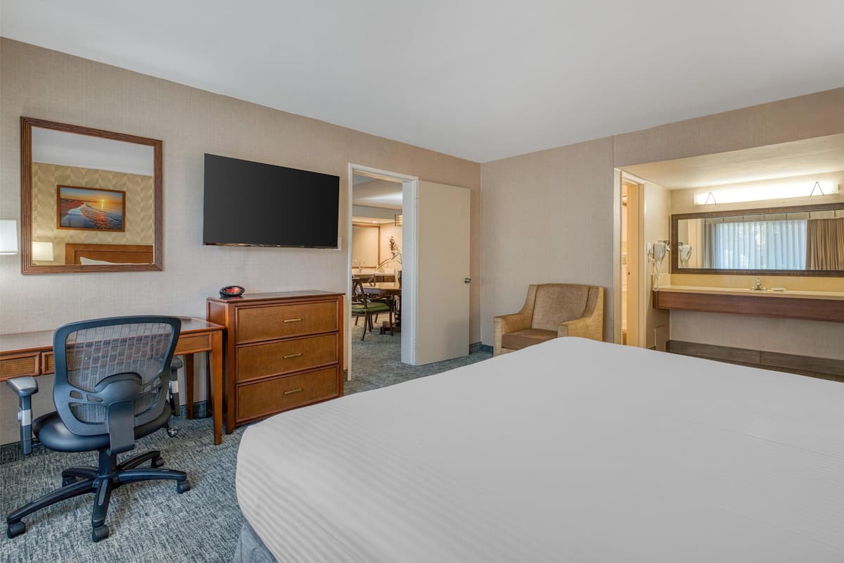 Ein Hotelzimmer mit einem großen Bett, einem Schreibtisch mit Computer, einem Stuhl, einem Schminktisch, einem Fernseher und einem Badezimmer mit Spiegel und Waschbecken.