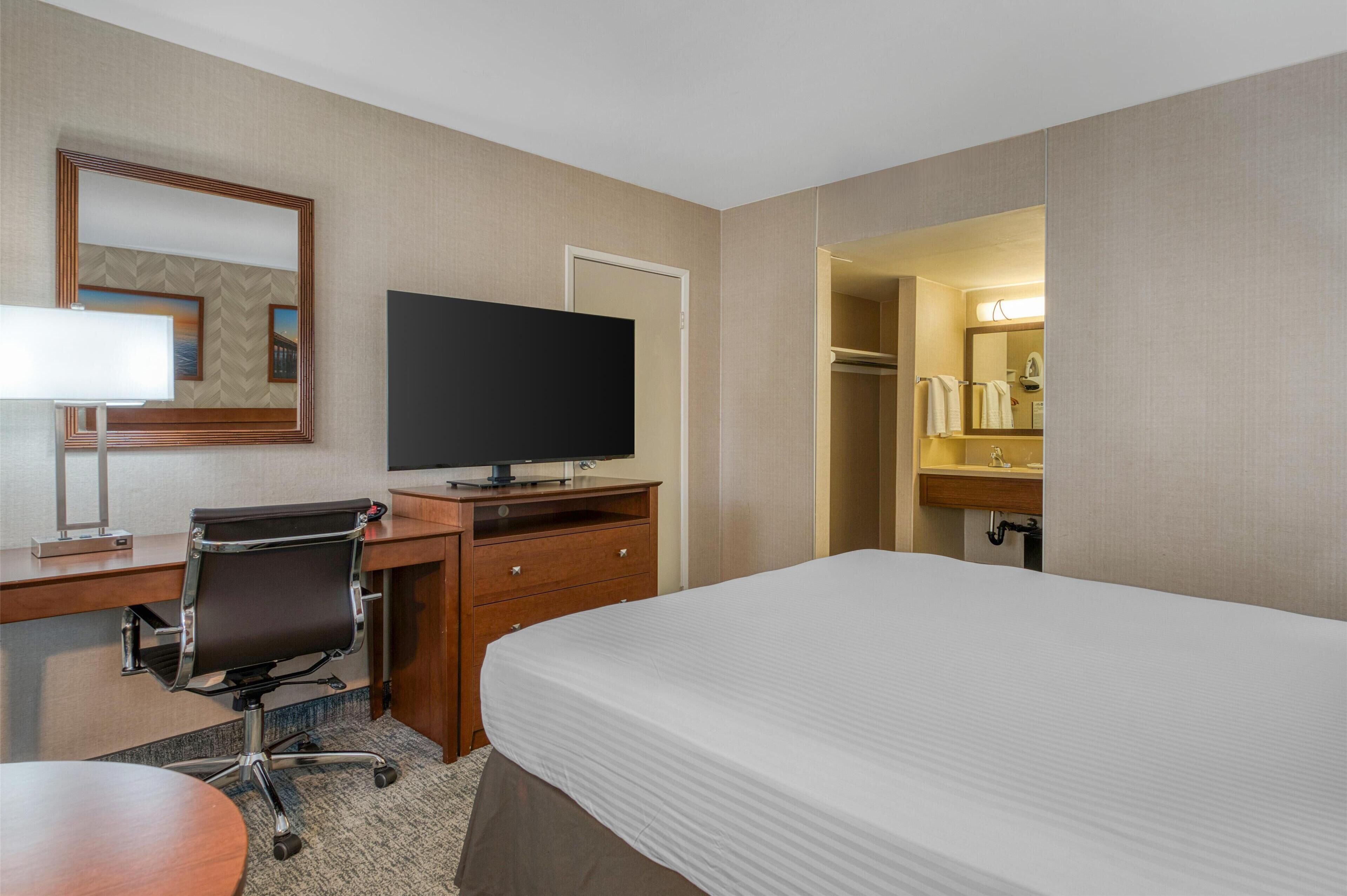 Quarto de hotel com cama, escrivaninha, cadeira, televisão e espelho.