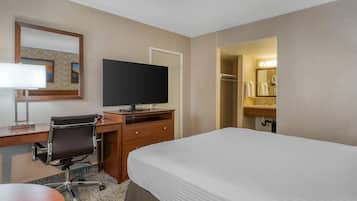 Quarto de hotel com cama, escrivaninha, cadeira, televisão e espelho.