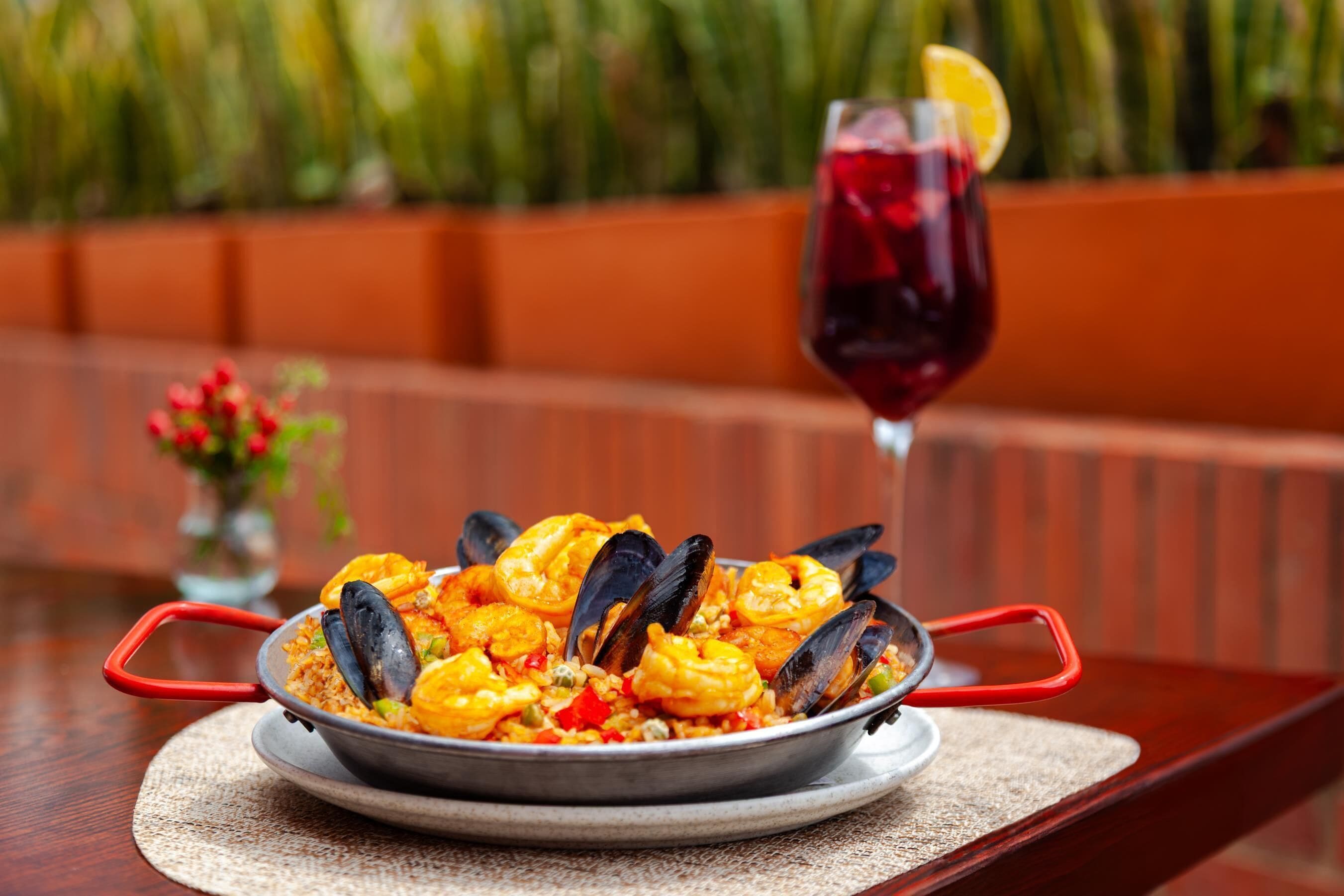 A seafood dish with mussels and shrimp, served on a metal plate with red handles, accompanied by a glass of red wine with a lemon slice.