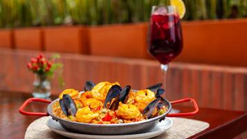 A seafood dish with mussels and shrimp, served on a metal plate with red handles, accompanied by a glass of red wine with a lemon slice.