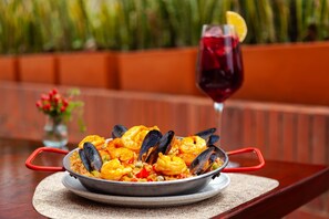 Un plato de mariscos con mejillones y camarones, servido en una fuente de metal con asas rojas, acompañado de una copa de vino tinto con una rodaja de limón.