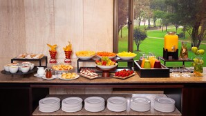 Un bufé con diferentes opciones de comida y bebidas, vista a un parque a través de una ventana, y platos ordenados.