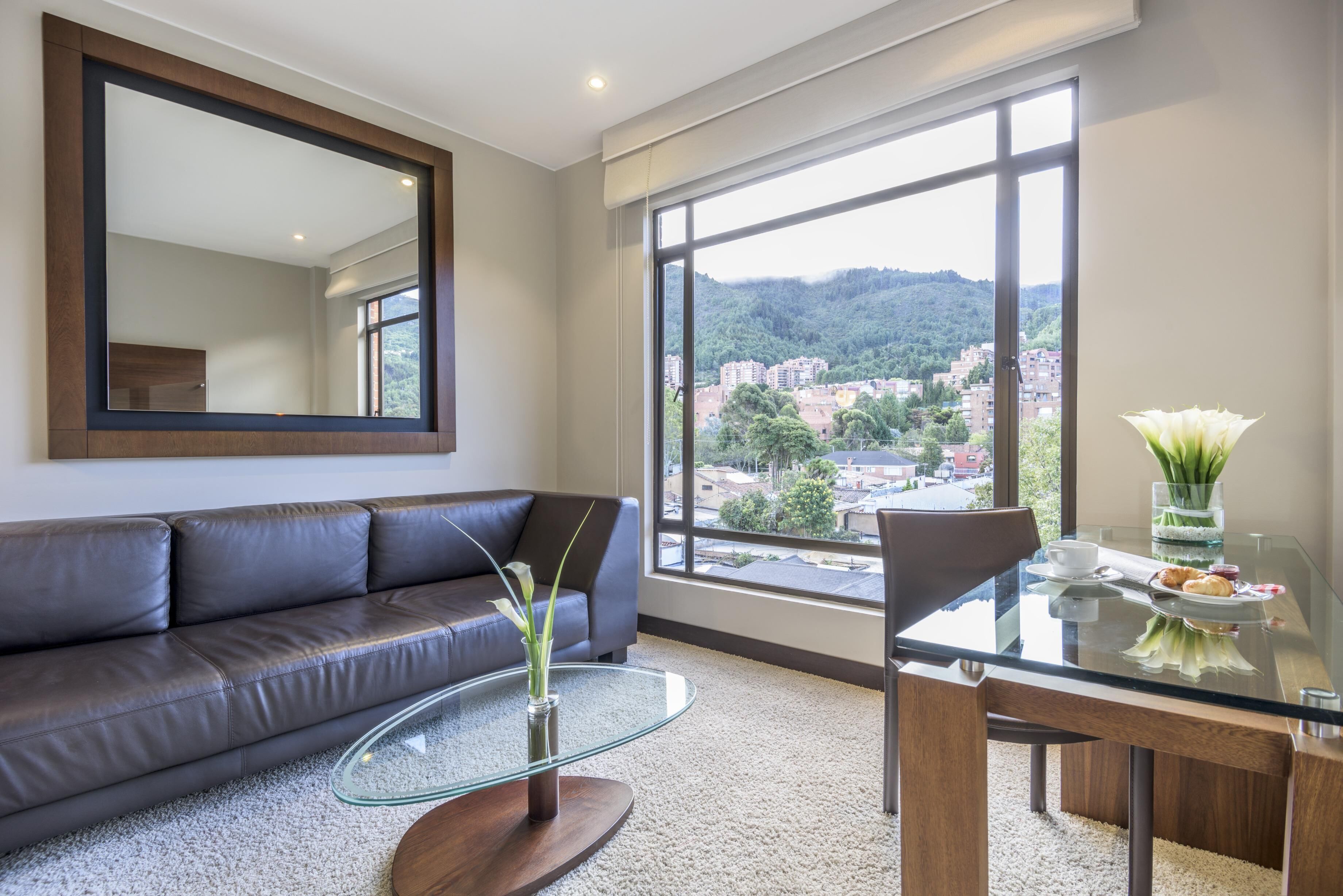 A modern living room with a leather sofa, a glass coffee table, and a dining table with chairs. A large window offers a view of a cityscape and hills.