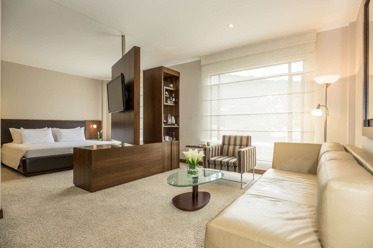 Habitación de hotel con cama, sofá, mesa de centro de vidrio, televisor montado en la pared y un ventanal grande.