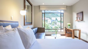 Habitación de hotel con una cama grande, un sofá, una mesita con un florero y vistas a un paisaje urbano a través de un ventanal amplio.