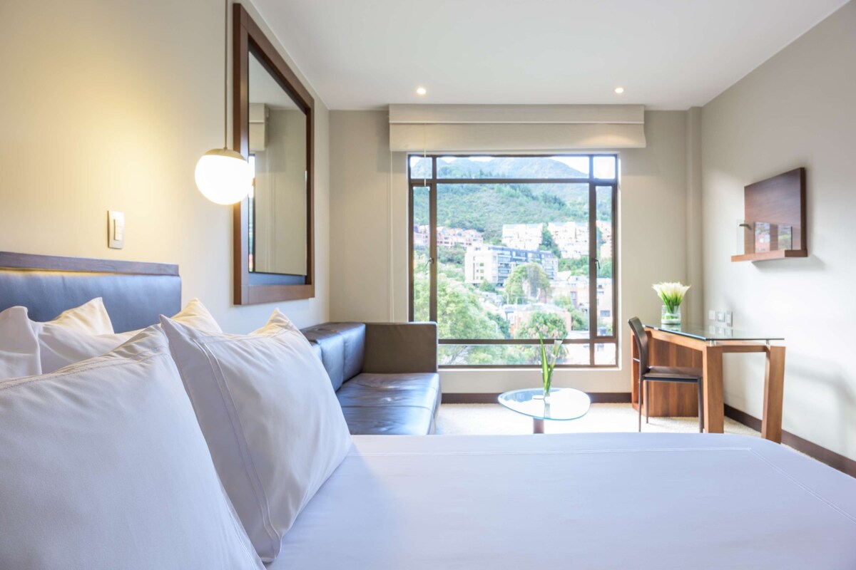 Habitación de hotel con cama, sofá, una mesita con un florero, una silla y un gran ventanal con vistas a edificios y árboles.