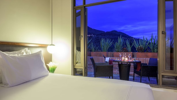 Habitación de hotel con una cama grande, zona de comedor con sillas y una mesa, y vista a una montaña al atardecer.
