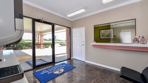 A reception area with a sliding glass door leading outside, a reception desk, a blue mat with a logo, a black office chair, and a framed picture on the wall. - Americas Best Value Inn Pasadena Houston (Pasadena)