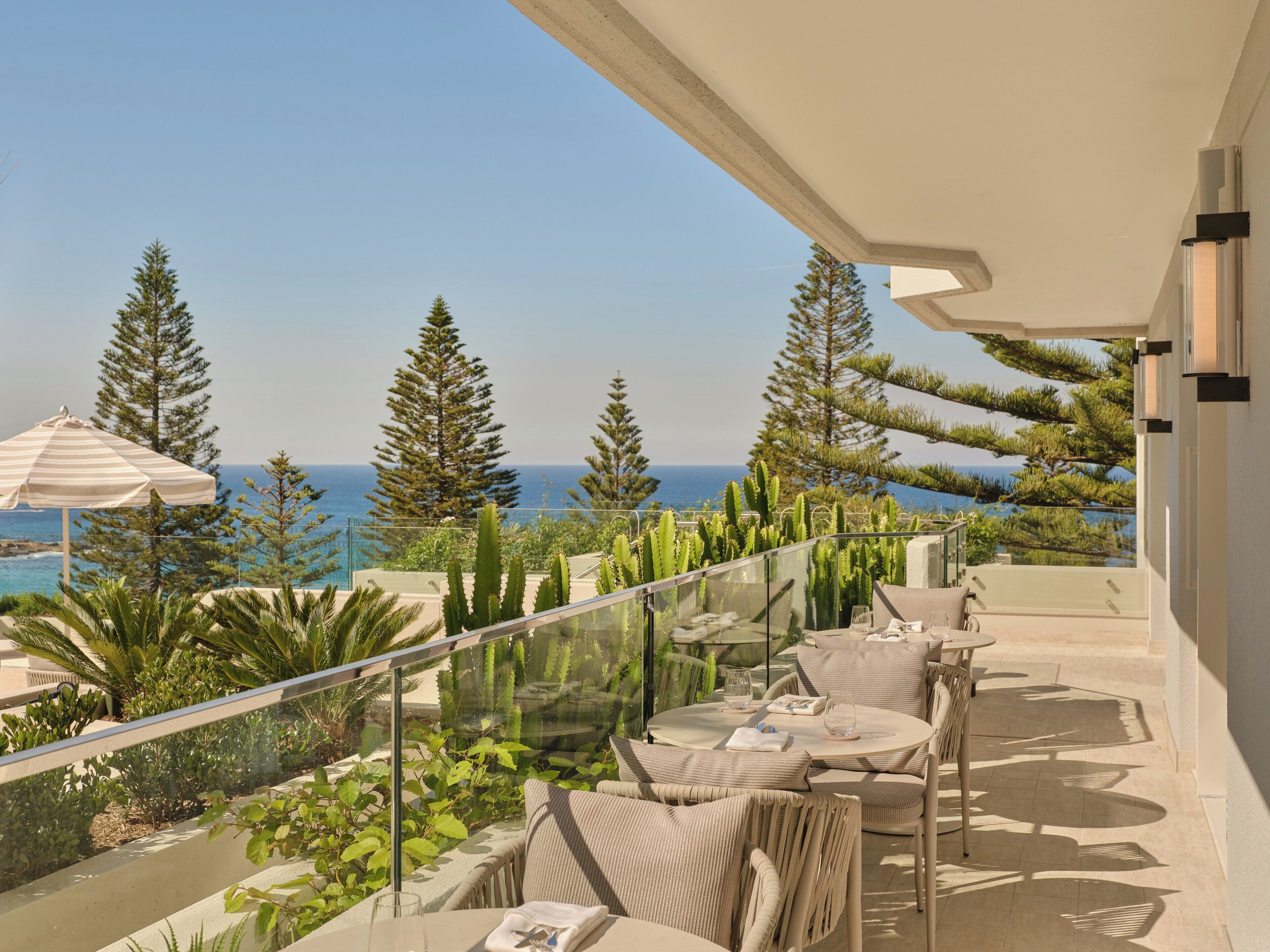 Un balcón con barandilla de vidrio, muebles de exterior y vista al mar y árboles.