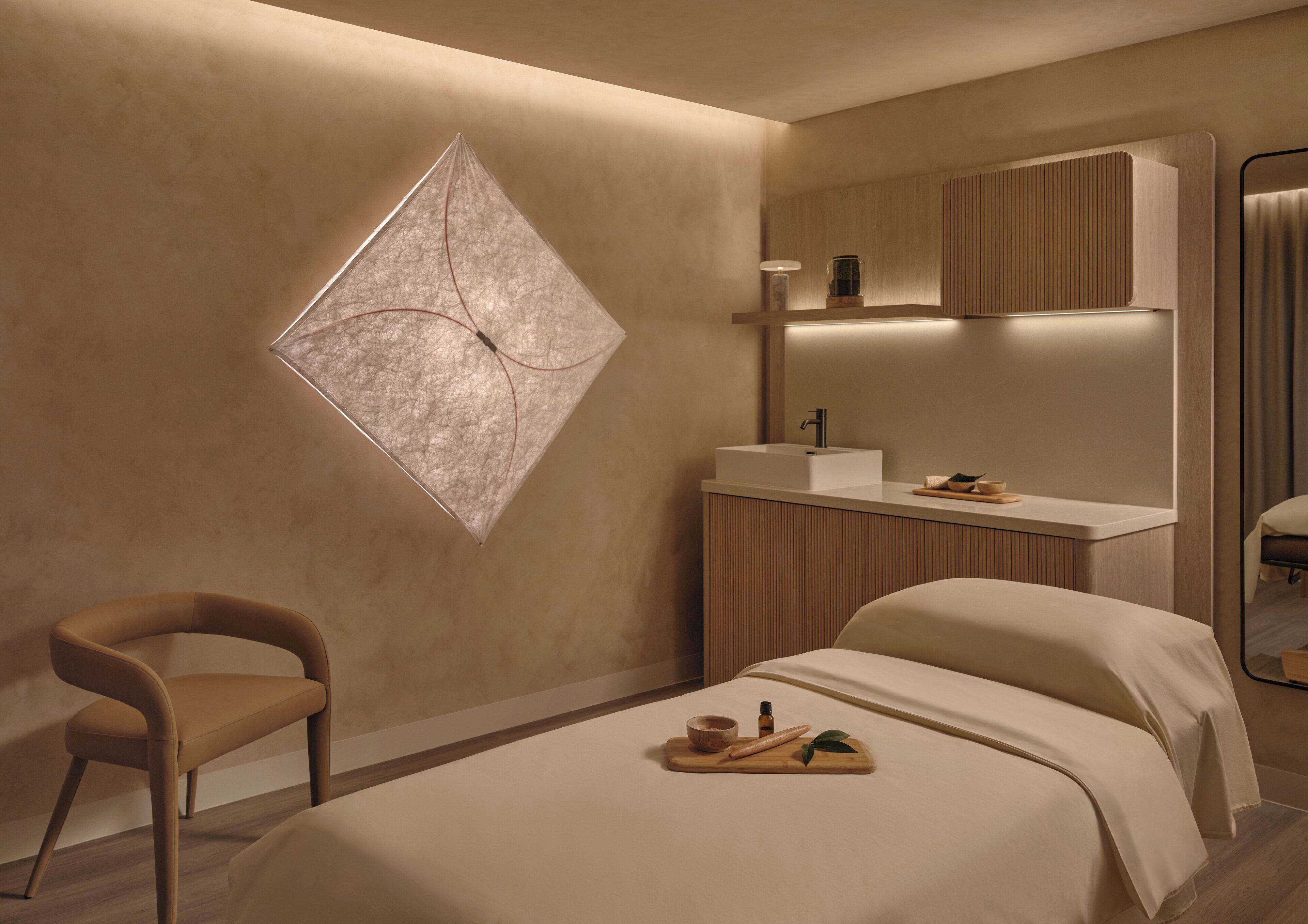 Una habitación de hotel moderna con una cama, una silla, una mesita de noche con lámpara, un espejo y una obra de arte colgada en la pared.