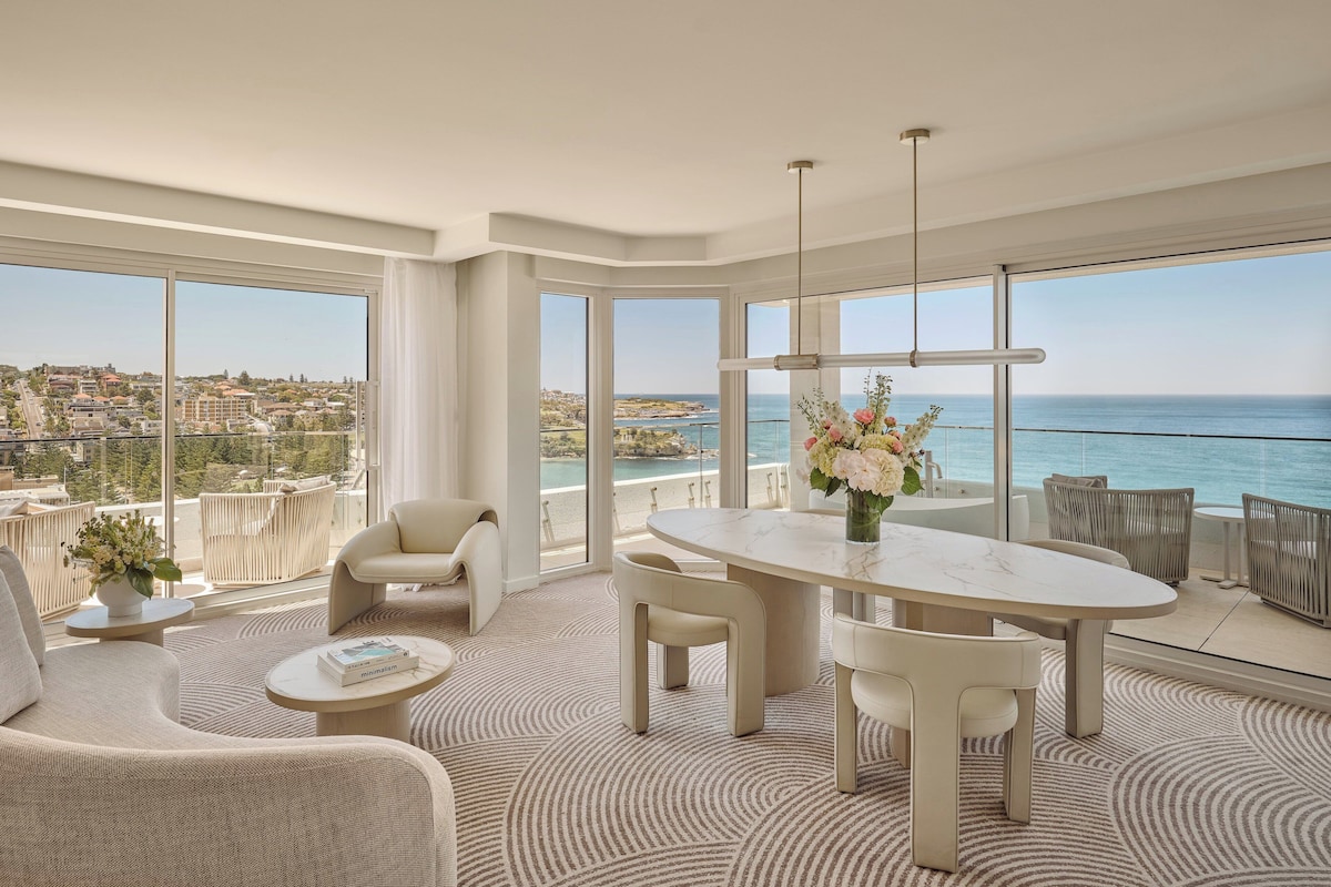 Un salon moderne avec une grande table ovale, des canapés de couleur beige et une vue sur l’océan.