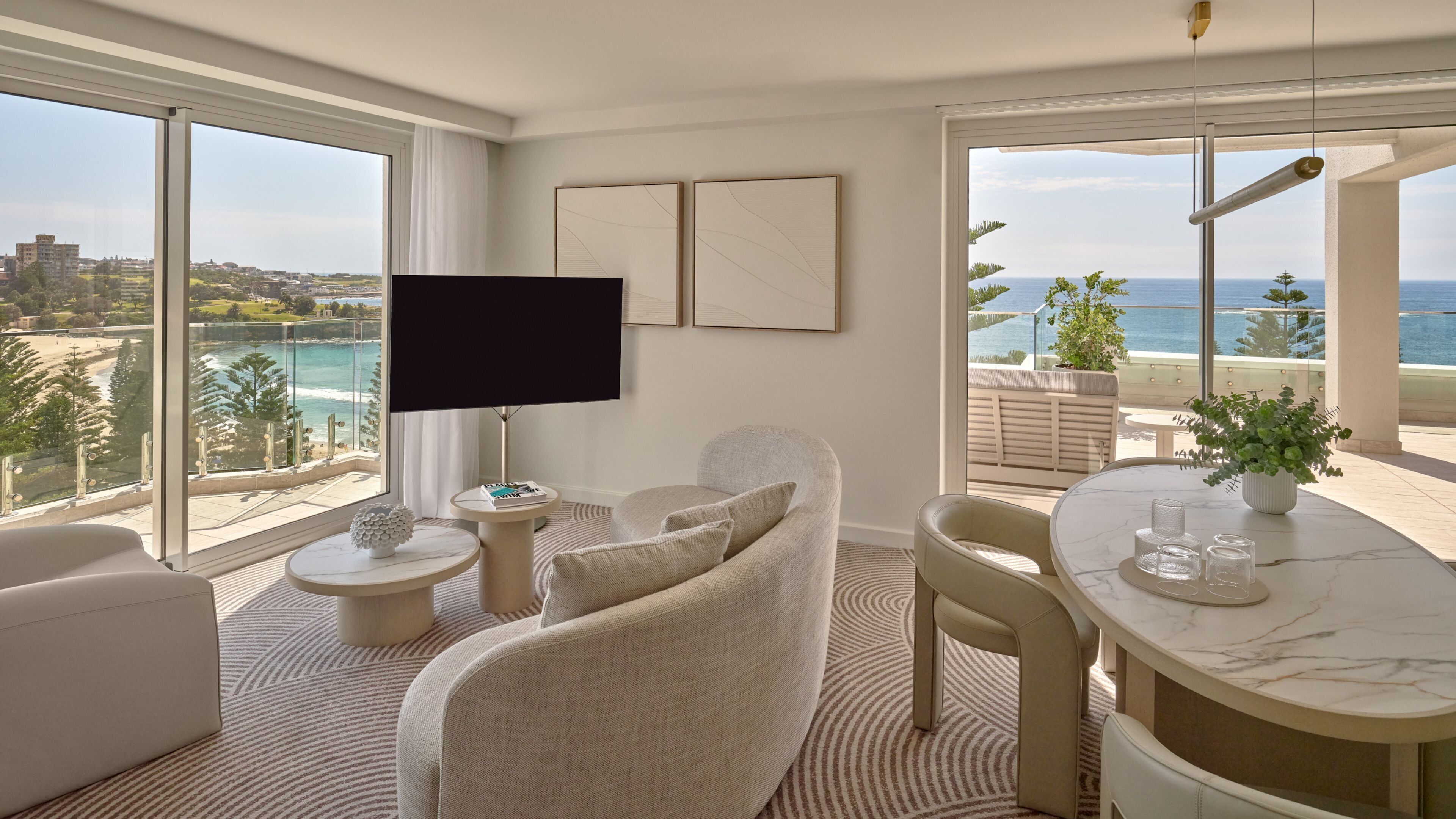 A modern living room with a large TV, comfortable seating, a dining table, and a view of the ocean.
