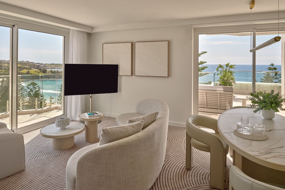 A modern living room with a large TV, comfortable seating, a dining table, and a view of the ocean.