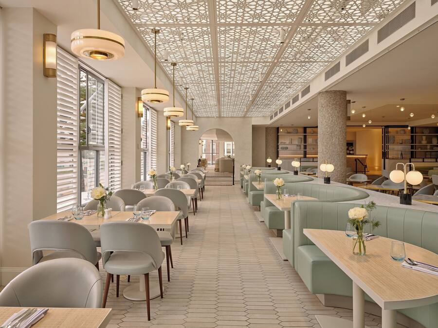 A modern restaurant with long tables and booths, a patterned ceiling, and a view of the street.