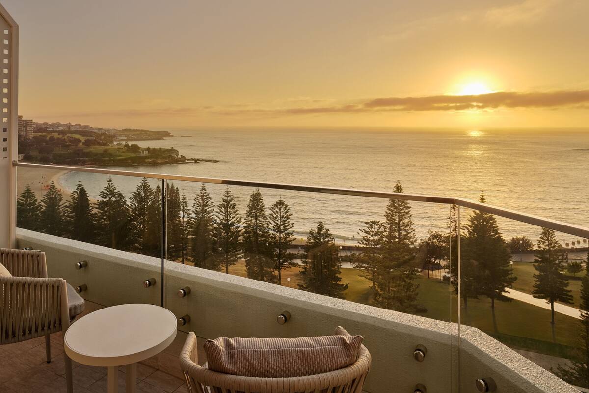 Un balcón con vista al mar al atardecer, con muebles de mimbre y barandilla de vidrio.