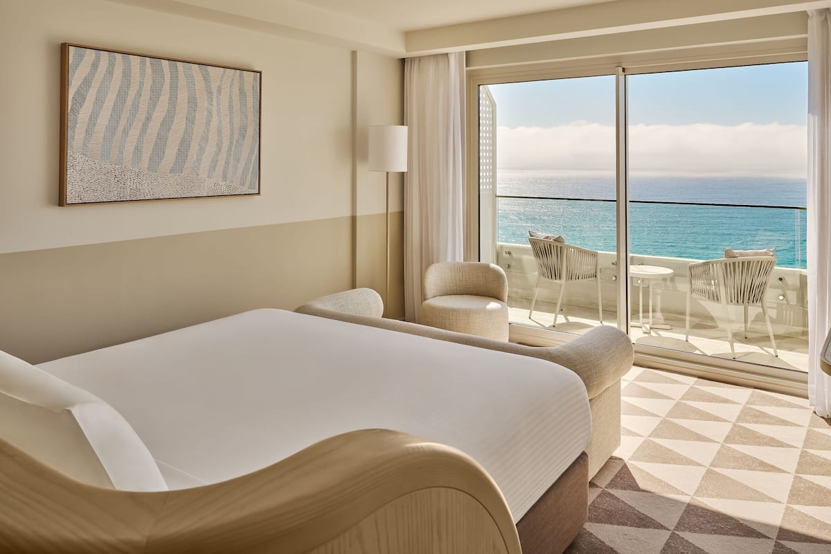Habitación de hotel con una cama grande, zona de estar con sillas, una mesa y balcón con vista al océano.