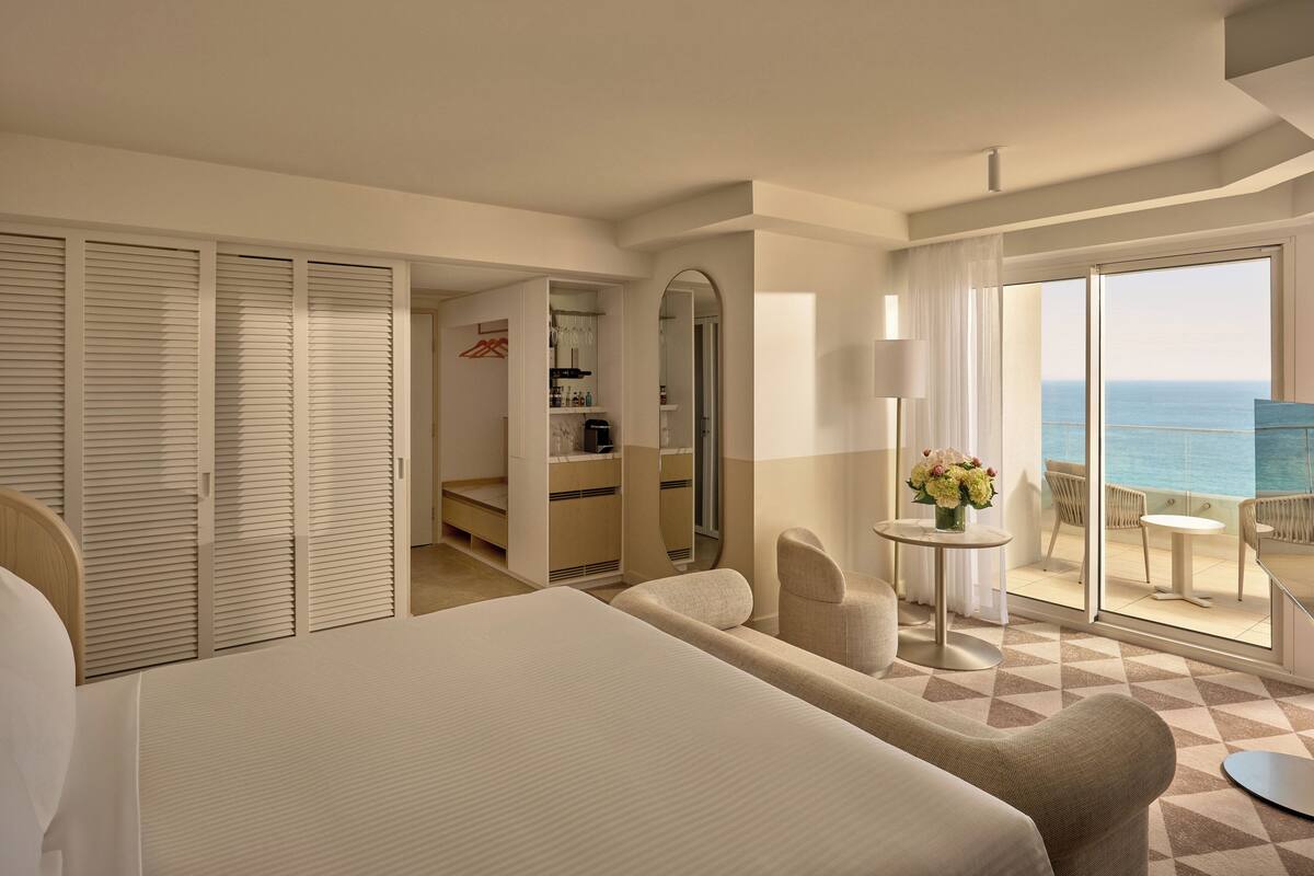 Habitación de hotel con una cama grande, zona de estar, una mesita con flores y balcón con vista al océano.