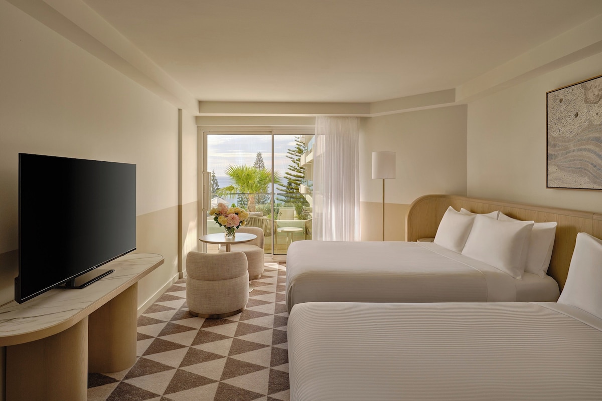 Une chambre d’hôtel avec deux lits, une télévision, une petite table avec des fleurs et un balcon aménagé avec du mobilier d’extérieur.