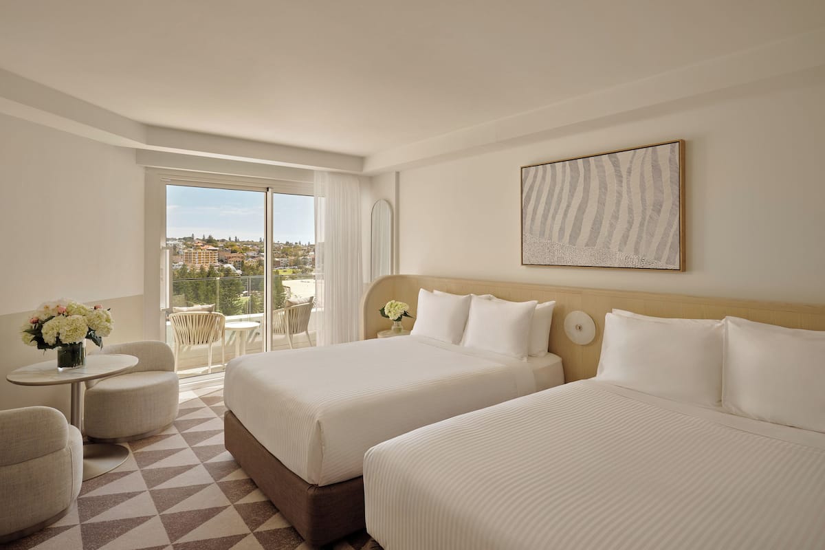 Ein Hotelzimmer mit zwei Betten, einem kleinen Tisch mit Blumen, einem Sessel und einem Balkon mit Blick auf die Stadt.