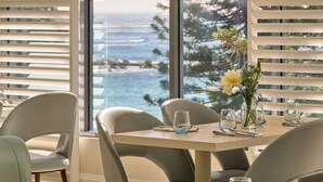 A dining area with a table set for four, large windows with blinds, and a view of the ocean.