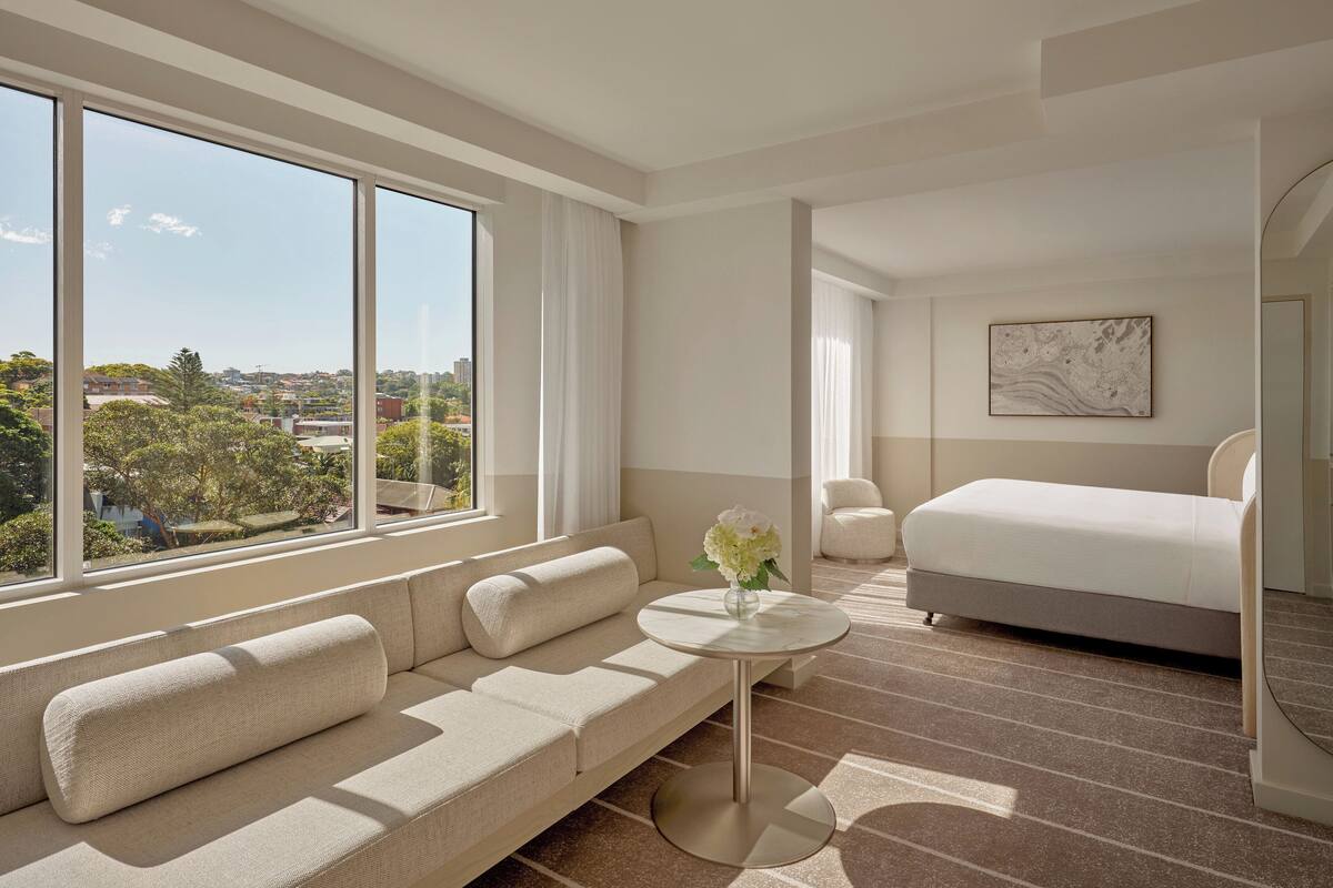 Una habitación de hotel moderna con un ventanal grande, un sofá, una cama, una mesita redonda y un cuadro en la pared.