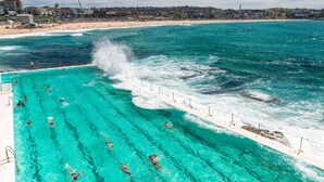 A swimming pool with clear blue water, a beach, and buildings in the background.