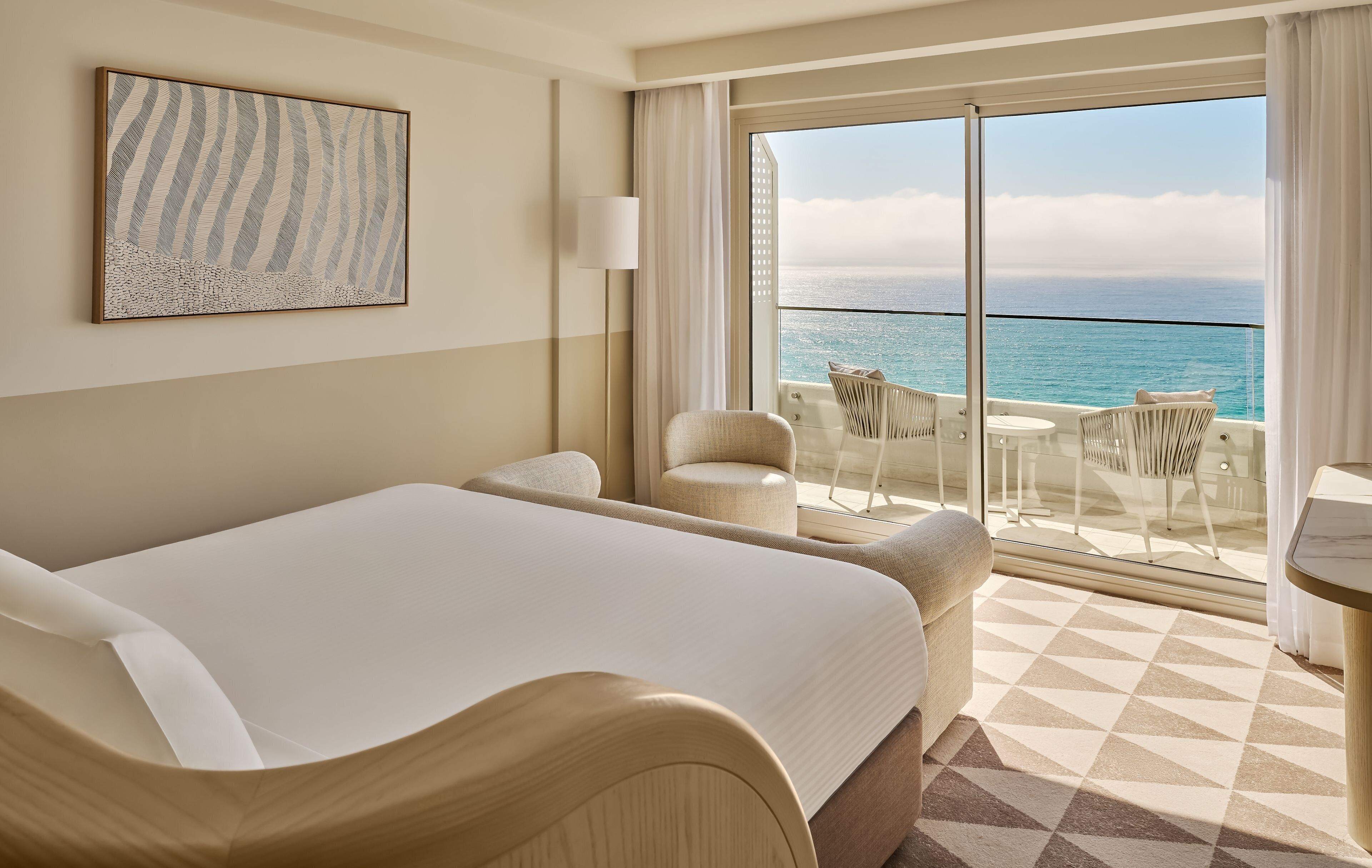 Une chambre d’hôtel dotée d’un grand lit, d’un coin salon avec des fauteuils, une table et un balcon offrant une vue sur l’océan.