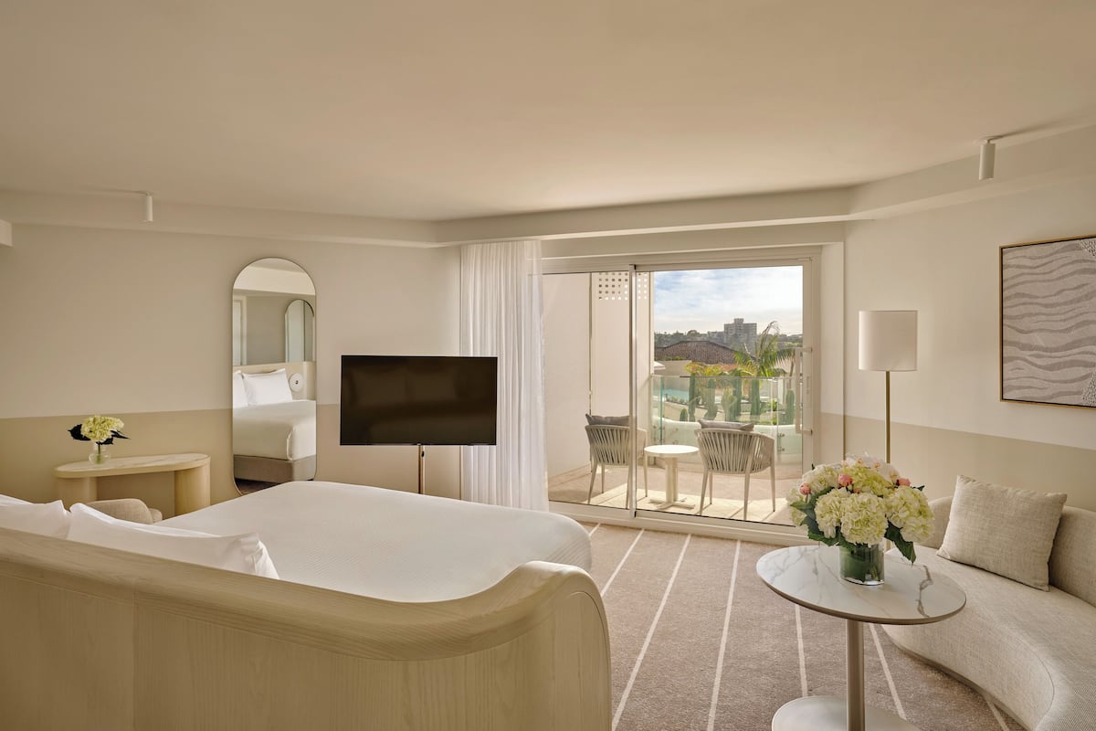 Habitación de hotel con cama, televisor, una pequeña mesa con un jarrón de flores, sofá y balcón con vistas a edificios.