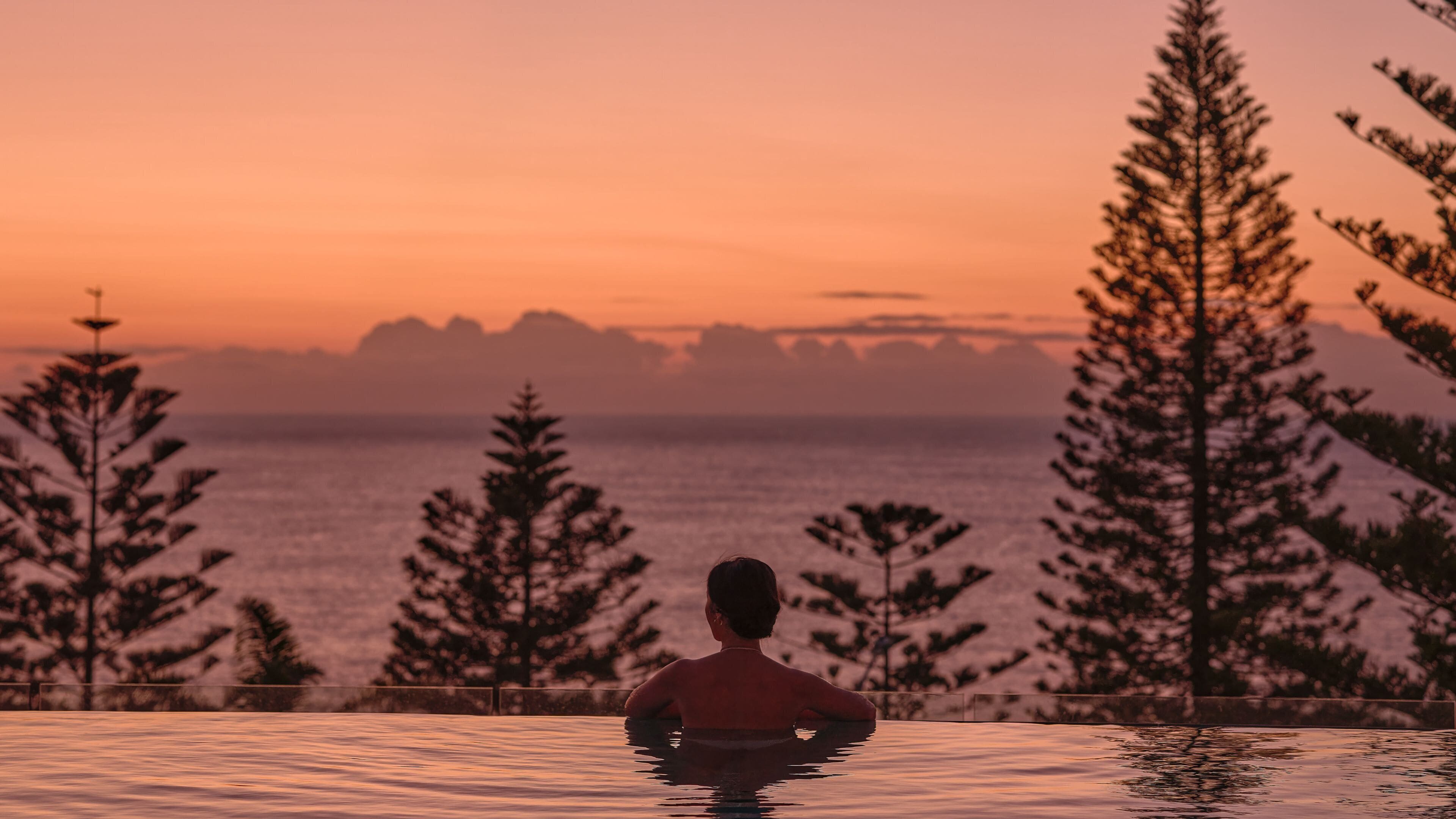 A person in a swimming pool with a view of the ocean and mountains during sunset.