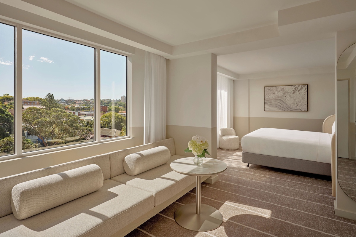 Une chambre d’hôtel moderne avec une grande fenêtre, un canapé, un lit, une petite table ronde et un tableau au mur.