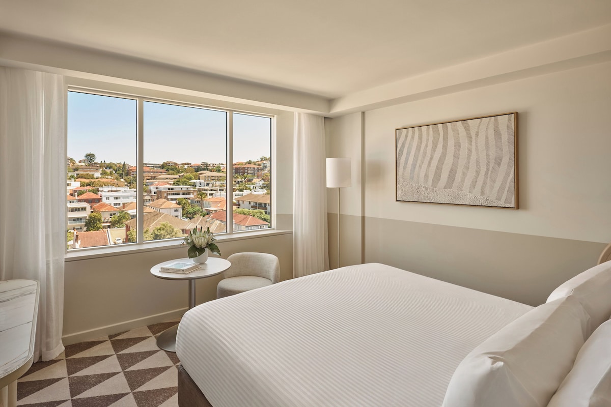 Une chambre d’hôtel avec un grand lit, une petite table ronde, une chaise et une fenêtre donnant sur la ville.