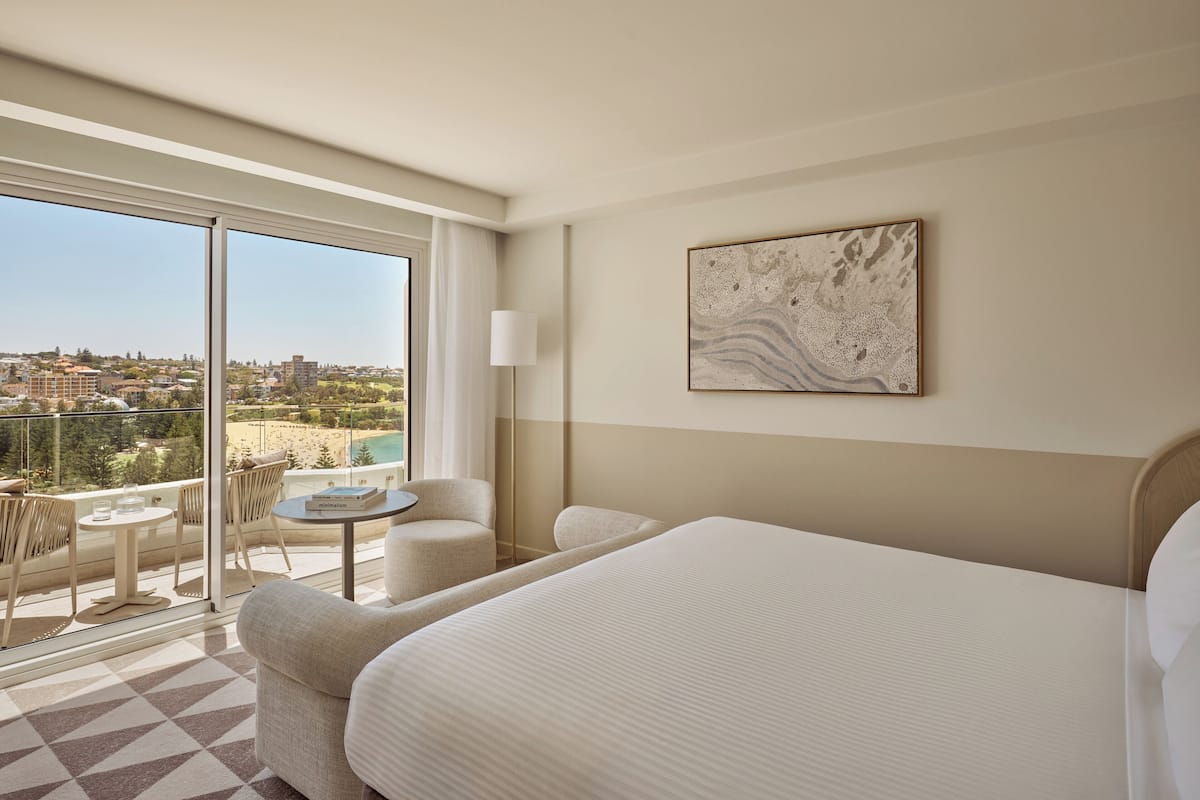 Ein Hotelzimmer mit einem großen Bett, einem kleinen runden Tisch mit einem Stuhl, einem Balkon mit Blick auf Strand und Stadt sowie einem an der Wand befestigten abstrakten Kunstwerk.