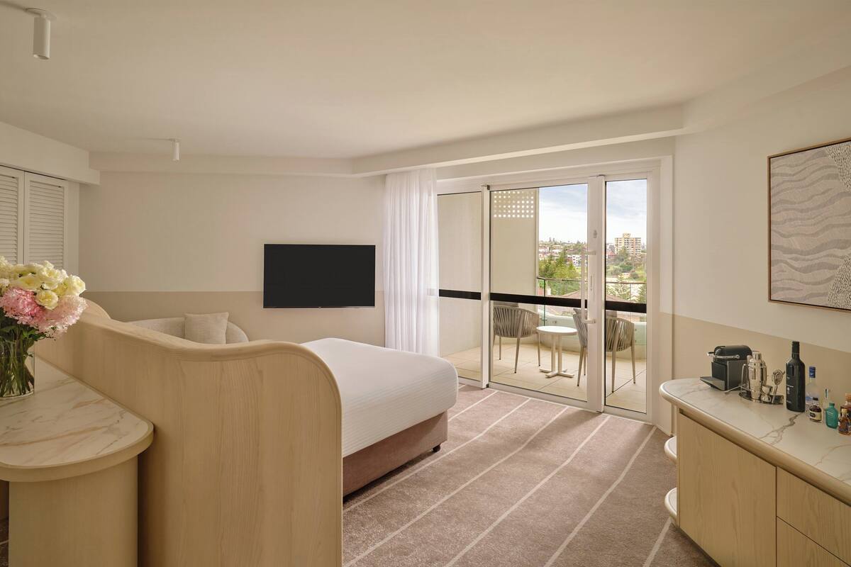 Una habitación de hotel con cama, escritorio, silla, televisión, balcón con vistas y una zona de bar.