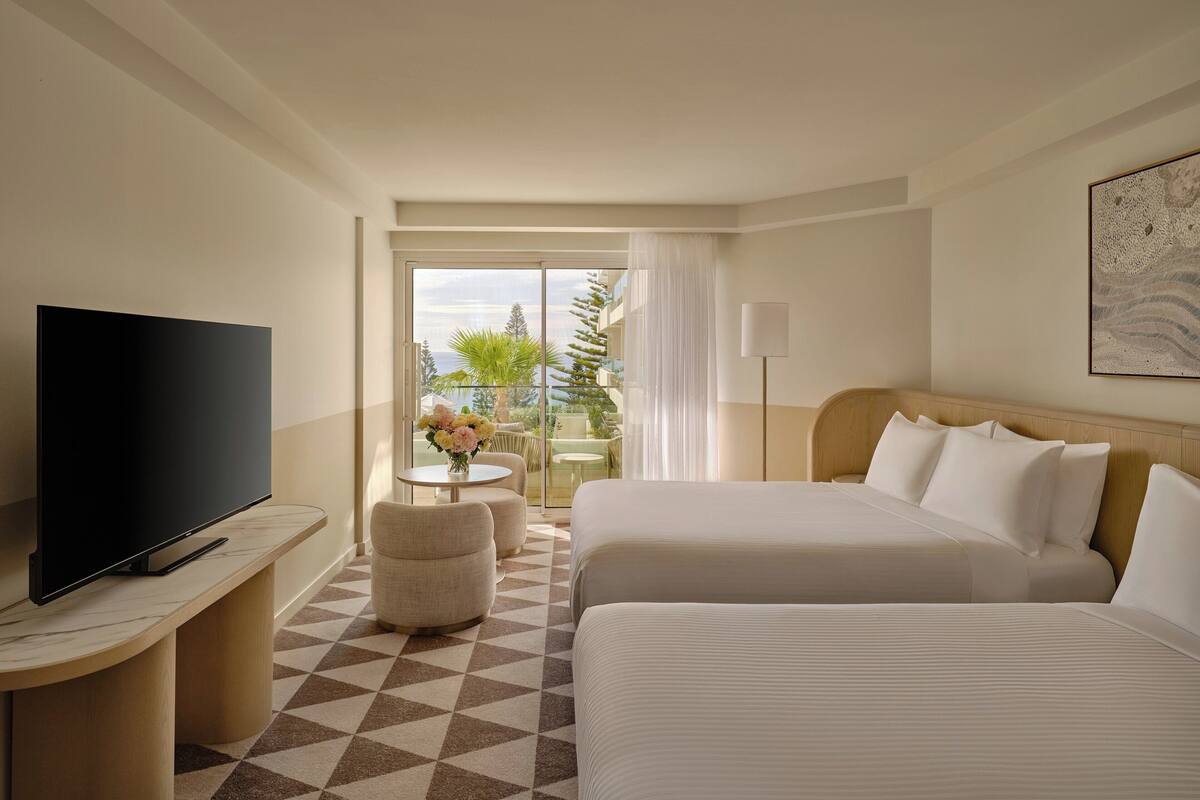 Habitación de hotel con dos camas, televisión, una pequeña mesa con flores y balcón con mobiliario exterior.