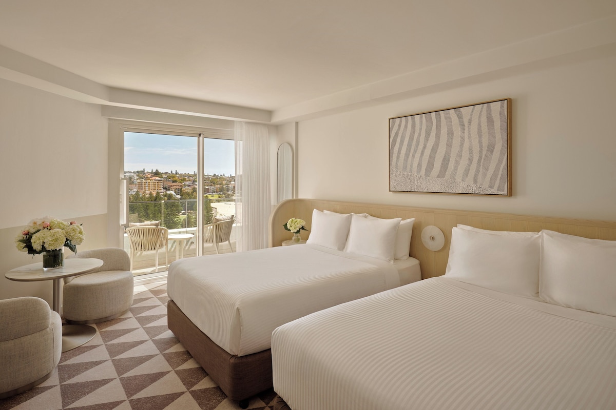 Une chambre d’hôtel avec deux lits, une petite table agrémentée de fleurs, une chaise et un balcon offrant une vue sur la ville.