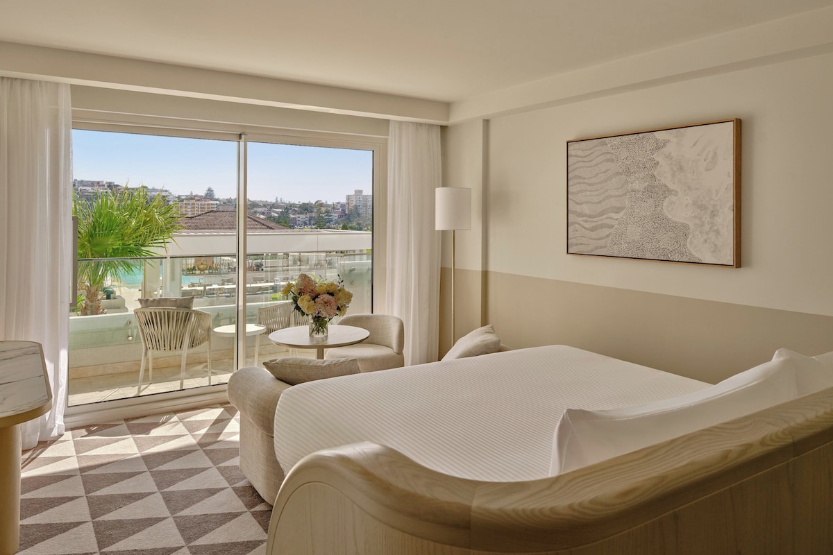 Une chambre d’hôtel avec un lit, une petite table ornée d’un vase de fleurs, une chaise et une vue sur la ville grâce à de grandes fenêtres.
