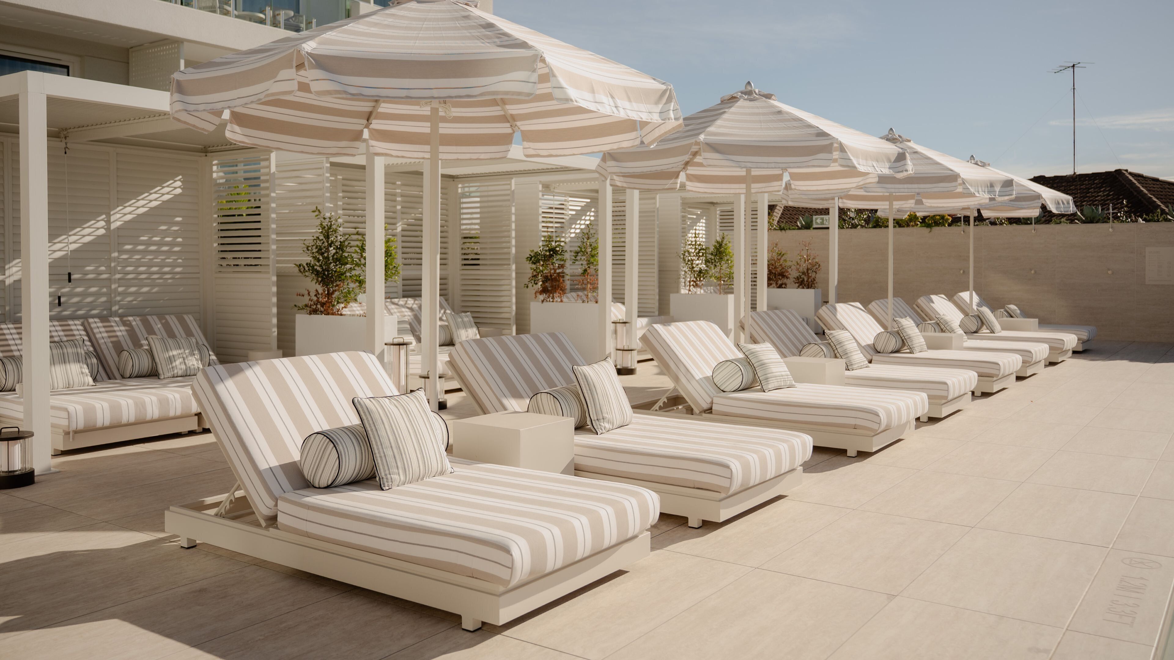 A row of sun loungers with striped cushions, white umbrellas, and potted plants.