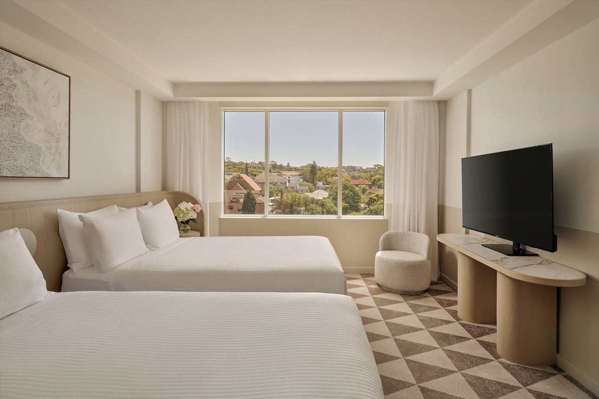 Habitación de hotel con dos camas, televisión, escritorio y ventana grande con vistas a edificios.
