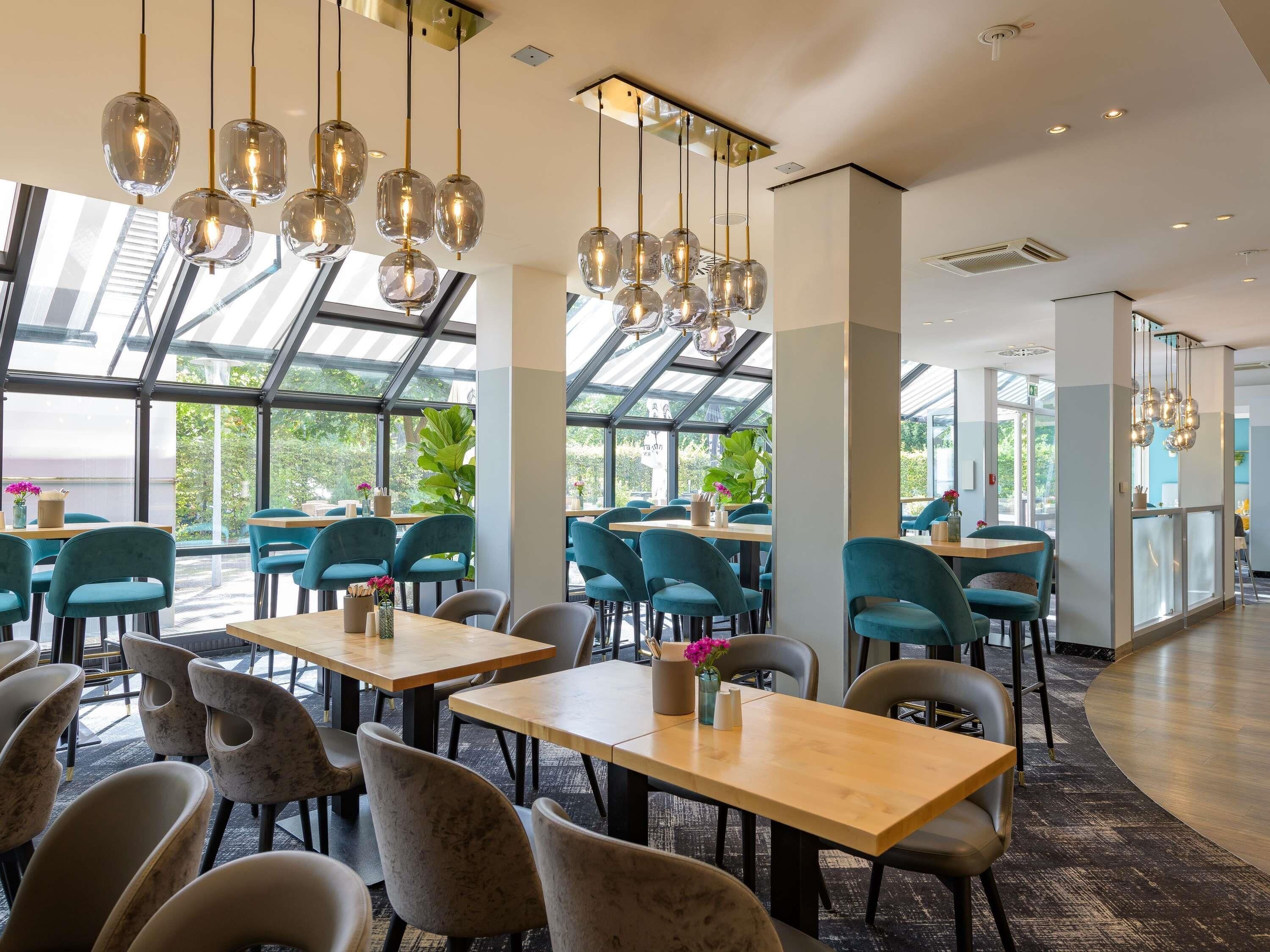 A modern restaurant interior with glass ceilings, teal chairs, wooden tables, and hanging pendant lights.