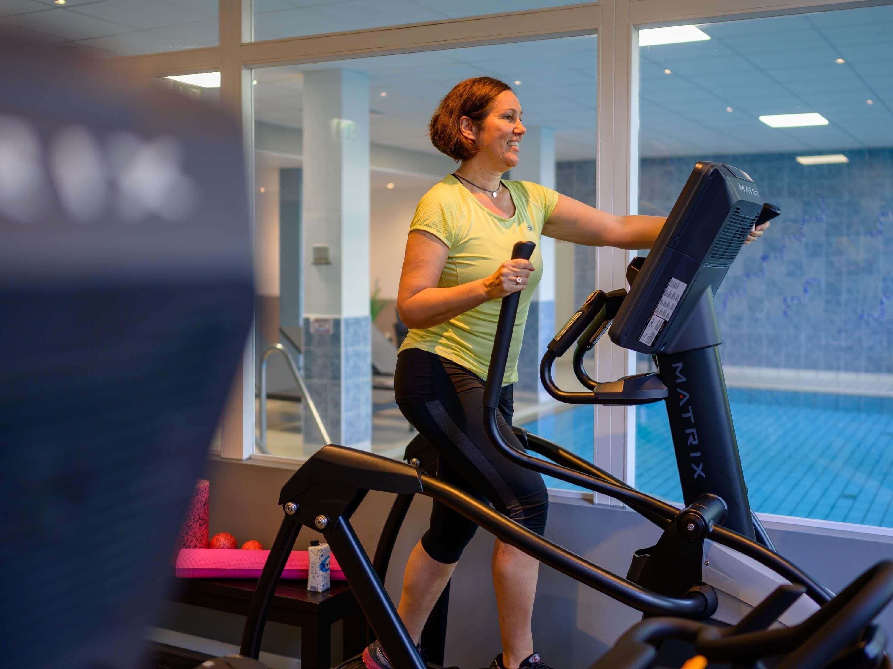 Una persona usando una cinta de correr en un gimnasio con un amplio ventanal y vista a una piscina.