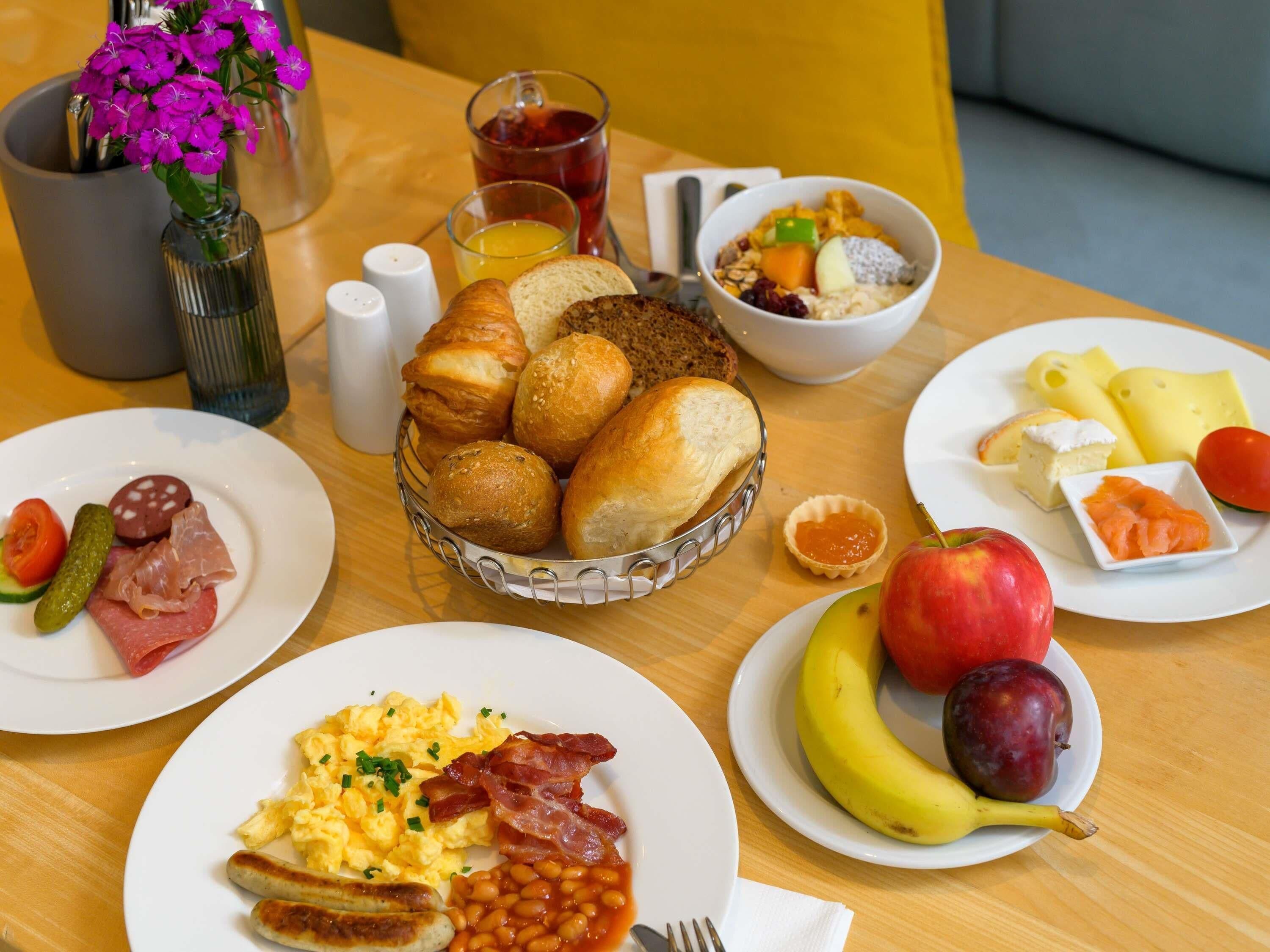 A breakfast setting with plates of food, a bowl of cereal, a vase of flowers, and a glass of juice.
