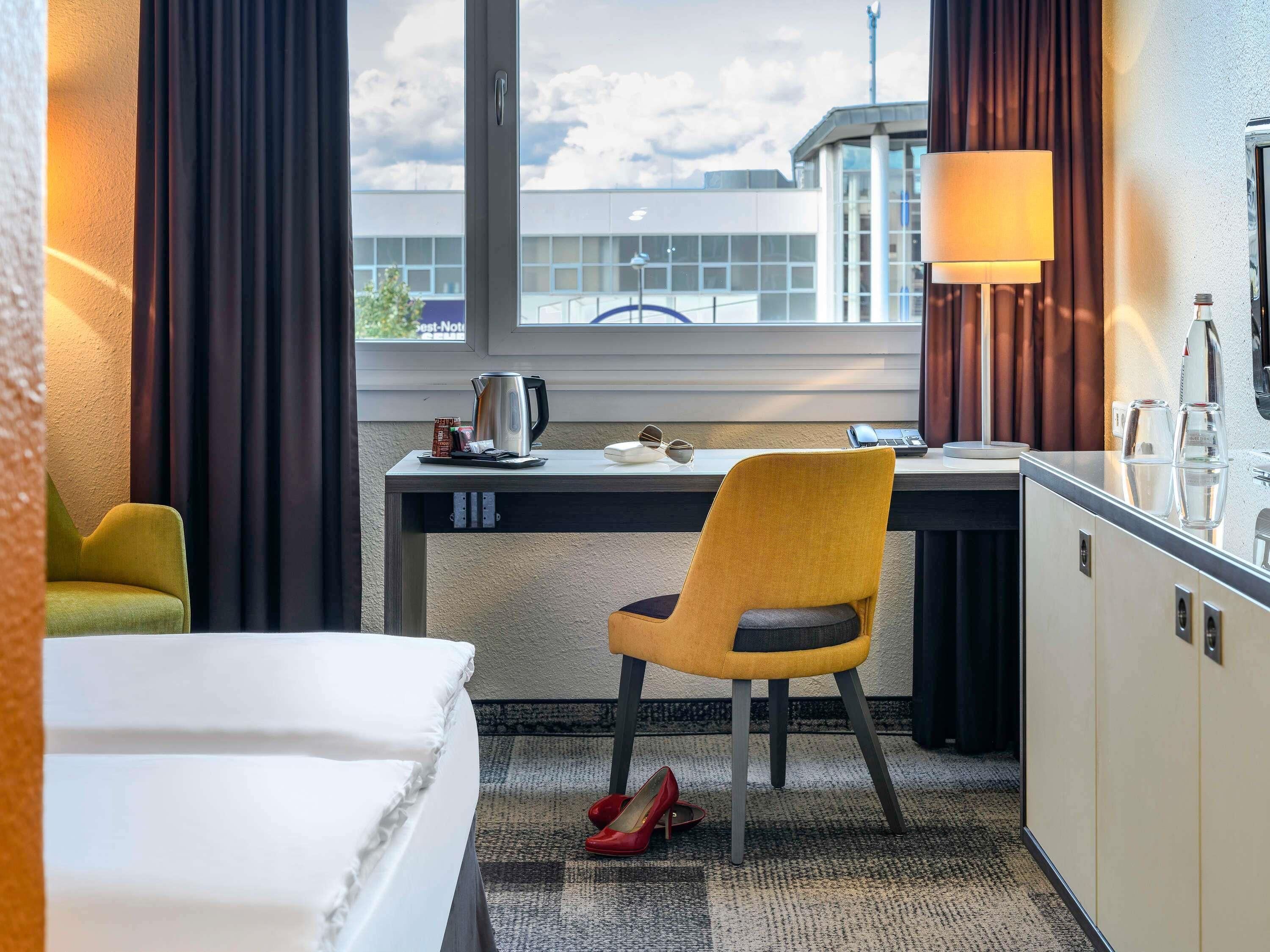 A hotel room with a bed, a desk, a chair, a lamp, and a view of the outside.