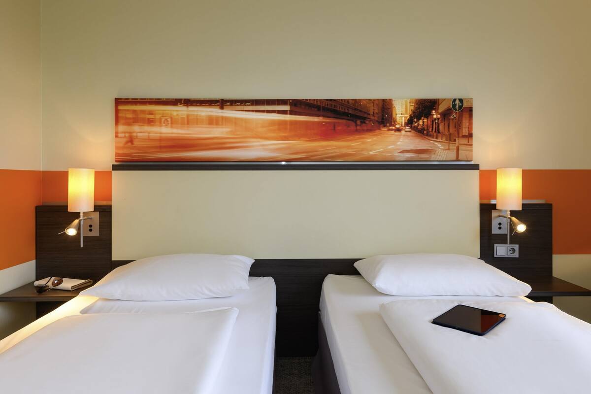 A hotel room with two single beds, a nightstand with a phone and a tablet, and a large painting of a city street on the wall.