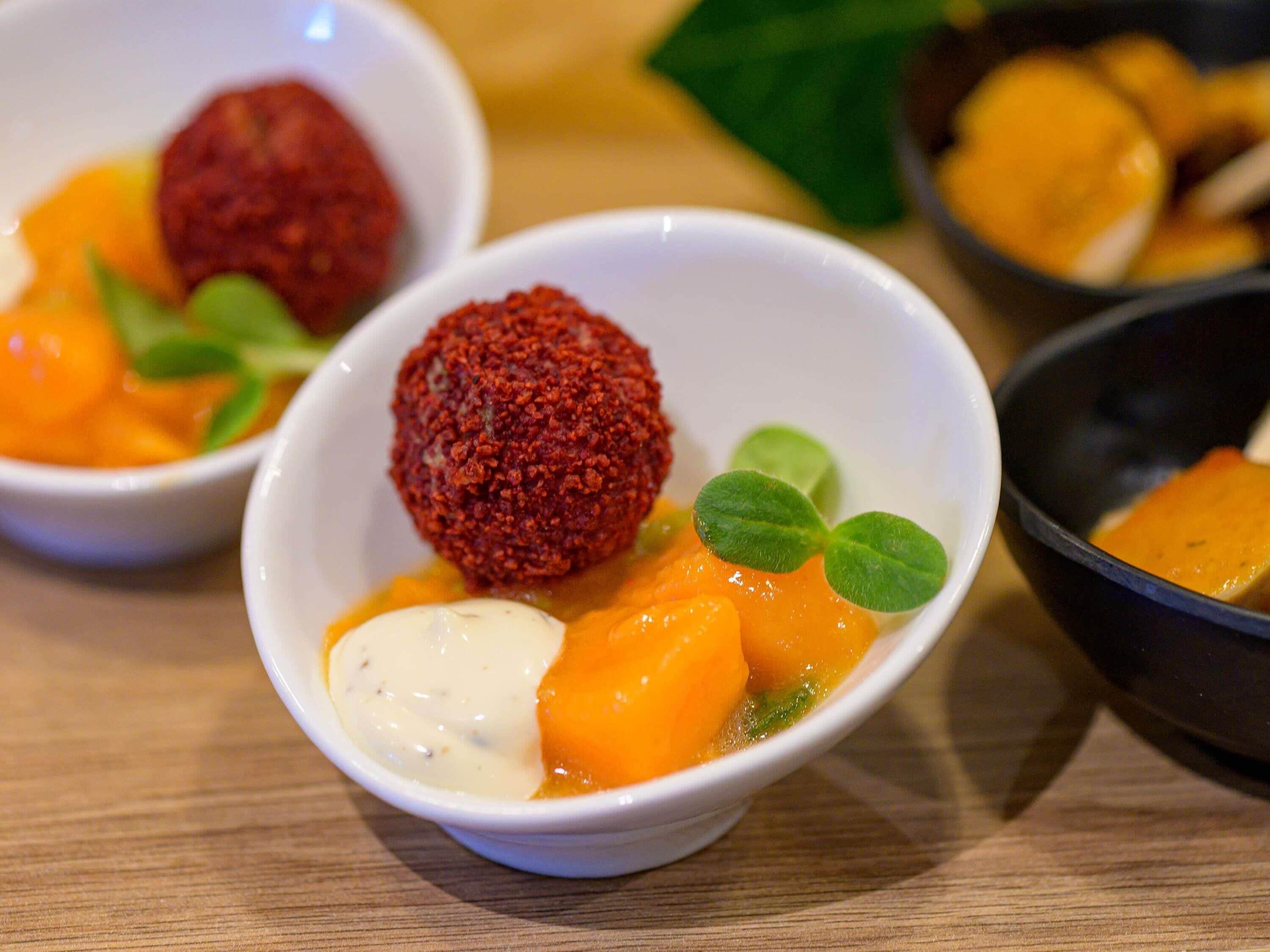 Three bowls of food with a red crumbly item, orange chunks, and a creamy sauce, on a wooden table.