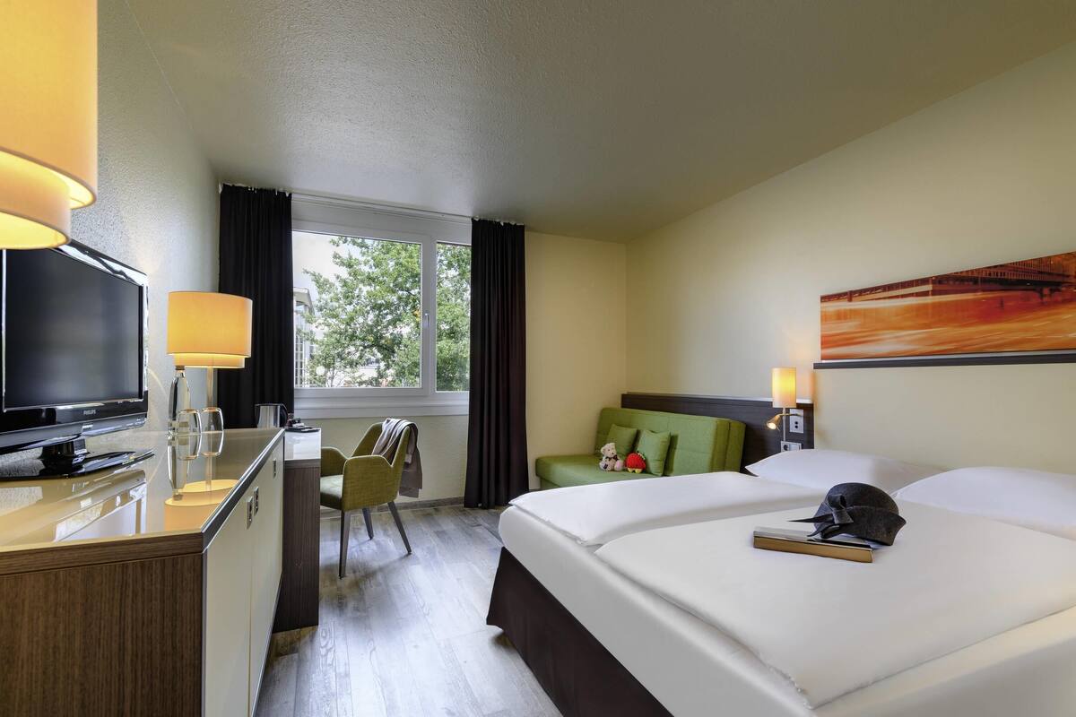 A hotel room with a bed, a desk with a television, a chair, a window with curtains, and a painting on the wall.