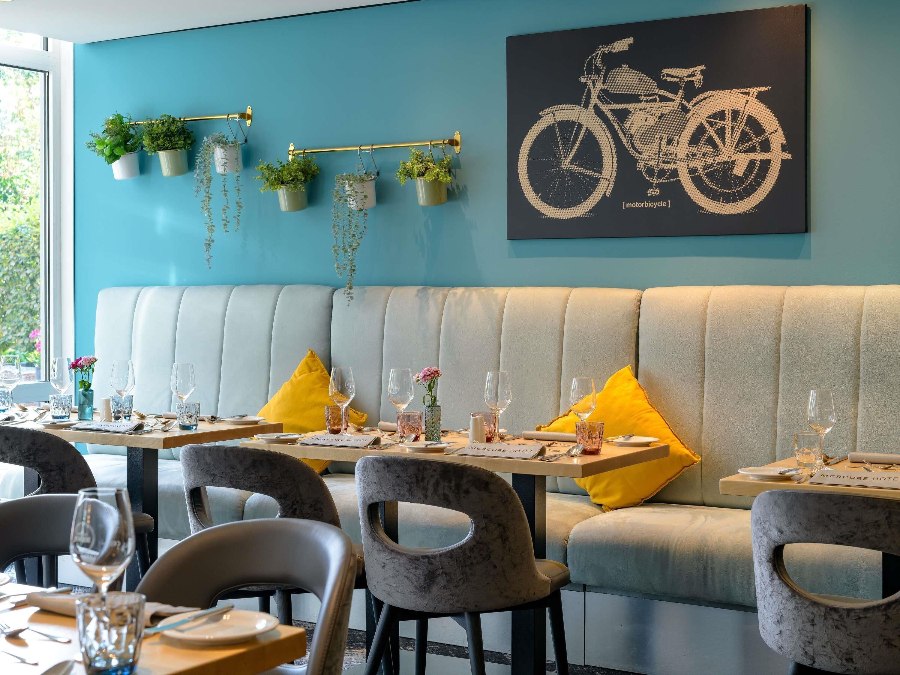 A restaurant with a teal wall, a motorcycle artwork, hanging plants, and a seating area with yellow cushions.