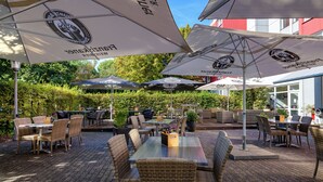 An outdoor patio area with wicker furniture, umbrellas, and a brick floor. - Mercure Hotel Köln West (Cologne)