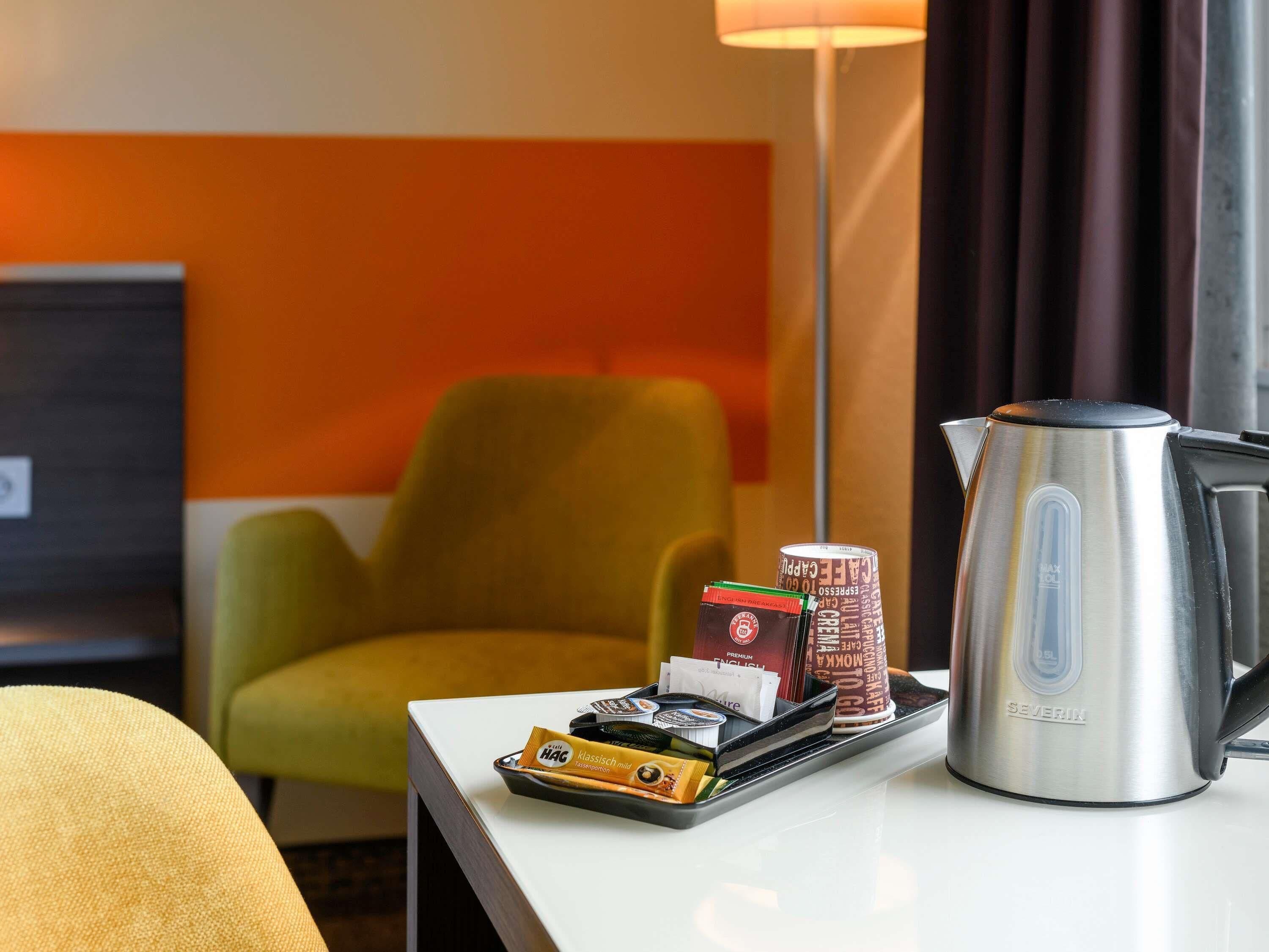 Une chambre d’hôtel avec une table blanche, un fauteuil vert, une bouilloire électrique argentée et un plateau contenant divers articles, notamment une boîte de thé et un thermos.