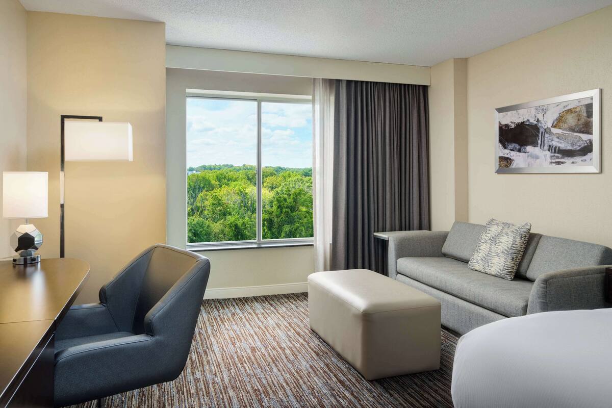 A hotel room with a large window, a sofa, a chair, a small table, and a bed.